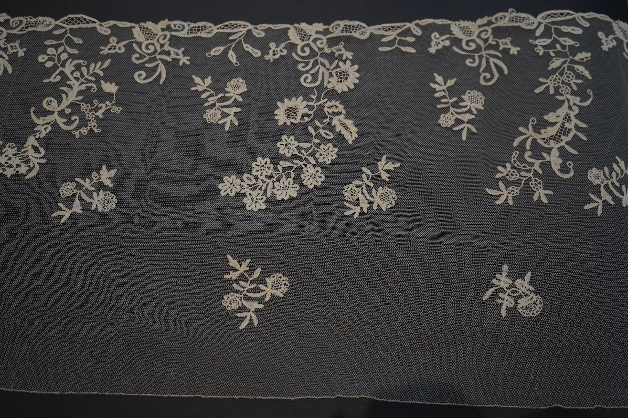 A 19th century five yard flounce of Brussels bobbin appliqué lace applied onto fine machine net, worked in a trailing floral design with an ornate border and sprig motifs, 457.5 cm long x 36cm deep. Condition - possibly